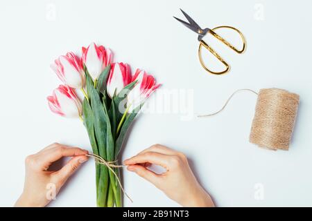Flat lay female's hands tying jute rope bow on simple bouquet of fresh tulips flowers on white table. Stock Photo