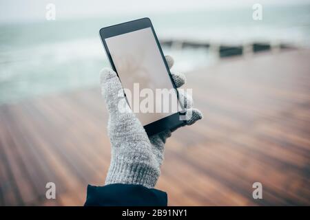 Female hand in glove holding mobile phone with blank display on background of wet wooden promenade and sea on winter day, close-up. Stock Photo