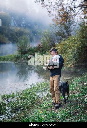 A man is holding a baby near a river and a dog Stock Photo