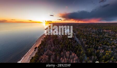 Roofs of houses in the forest, the picturesque coast at sunset, quiet water, tops of trees are illuminated by the sunset sun, pink clouds Stock Photo