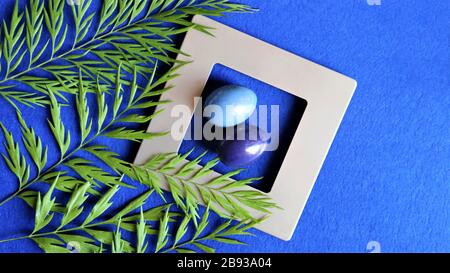 On a blue trendy background, eggs in a gray, silver frame. Green leaves of tropical plants. Trendy color background. Close up. Copy space Stock Photo