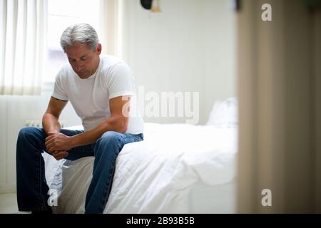 Contemplative mature man sitting on the edge of the bed in his bedroom. Stock Photo