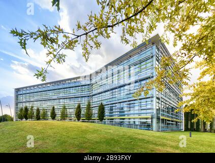 Exterior of a modern angular glass fronted office building in a spring landscape with green lawns and trees under a blue cloudy sky Stock Photo
