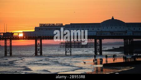 Brighton UK 23rd March 2020 - Walkers and a dog enjoy a stroll along  the beach as the sun sets behind the Brighton Palace Pier this evening after a beautiful sunny Spring day during the Coronavirus COVID-19 pandemic crisis . Credit: Simon Dack / Alamy Live News Stock Photo