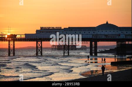 Brighton UK 23rd March 2020 - Walkers enjoy a stroll along  the beach as the sun sets behind the Brighton Palace Pier this evening after a beautiful sunny Spring day during the Coronavirus COVID-19 pandemic crisis . Credit: Simon Dack / Alamy Live News Stock Photo
