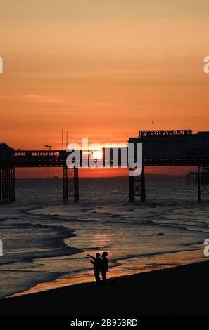 Brighton UK 23rd March 2020 - Walkers enjoy a stroll along  the beach as the sun sets behind the Brighton Palace Pier this evening after a beautiful sunny Spring day during the Coronavirus COVID-19 pandemic crisis . Credit: Simon Dack / Alamy Live News Stock Photo