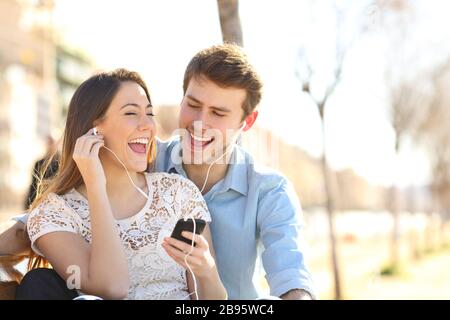 Happy couple singing together listening to music from smart phone sharing earphones sitting on a bench in a park Stock Photo