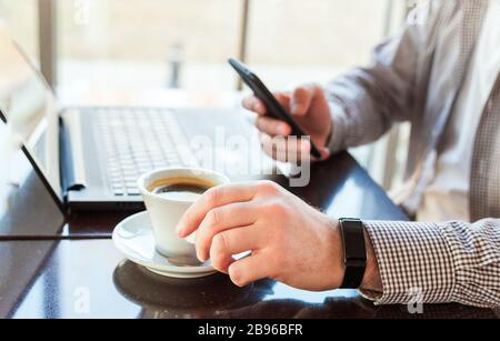 Man in smart watch drinking coffee on work space. Man using laptop, holding smart phone for business work or study. Close up smart watch on male hand Stock Photo