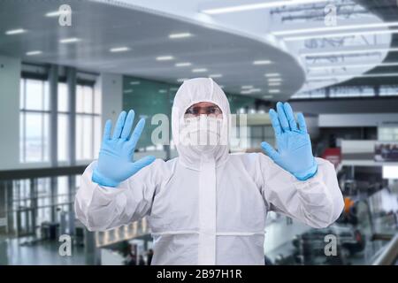 man epidemiologist in coverall disposable anti-epidemic antibacterial isolation suit shows a prohibition gesture against the interior of the airport o Stock Photo