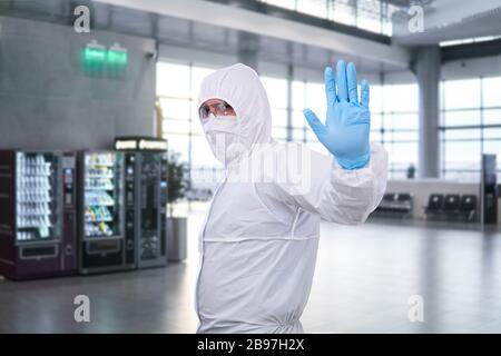 male epidemiologist in coverall disposable anti-epidemic antibacterial isolation suit shows a prohibition gesture against the waiting room at the airp Stock Photo