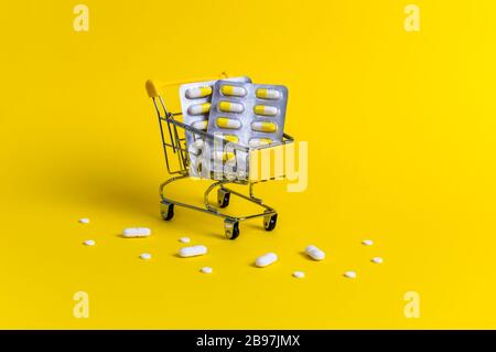 Shopping cart with blister packs and surrounded with white pills on yellow background. Creative concept of healthcare and safe online shopping for med Stock Photo