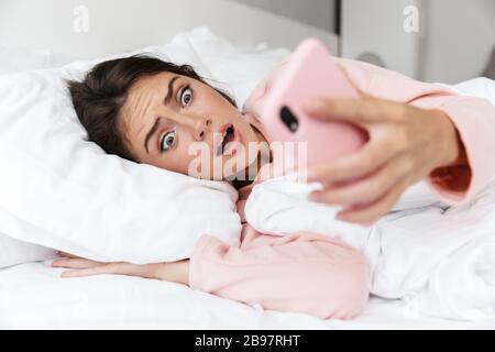 Shocked young girl wearing pajamas laying in bed at the bedroom, looking at mobile phone Stock Photo