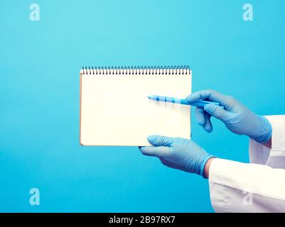 medic woman in white coat, blue latex gloves holding an open notebook with blank white sheets, blue background, place for text Stock Photo
