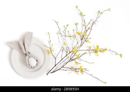 Easter festive spring table setting decoration. An egg on a gray napkin in the shape of a rabbit and a yellow forsythia branch on a white background. Stock Photo