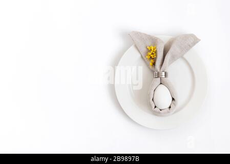 Happy easter concept. Easter table setting with egg in gray napkin Easter Bunny and a branch of yellow forsythia flowers on a white background. Stock Photo