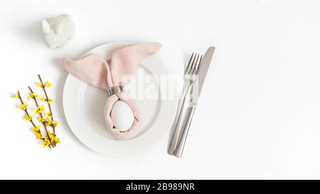 Easter holiday table setting with egg in pink napkin Easter Bunny and yellow forsythia flowers on a white background. Copy space, top view, flat lay. Stock Photo