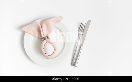 Easter table setting with egg in pink Easter bunny napkin on a white background. Copy space, top view, flat lay. Stock Photo