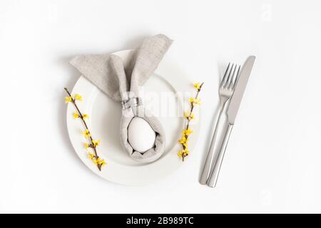 Easter holiday table setting with egg in gray napkin Easter Bunny and yellow forsythia flowers on a white background. Close up, top view, flat lay. Stock Photo