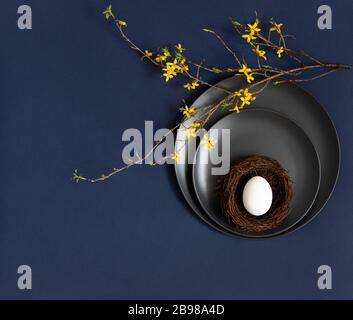 Easter table setting with a chicken egg in a nest on black plates and yellow flowers on a classic blue background. Copy space, top view, flat lay. Stock Photo