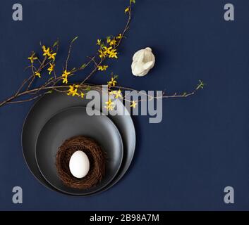 Easter table setting with a white egg in a nest , a porcelain rabbit and yellow forsythia flowers on a classic blue background. Copy space, top view Stock Photo
