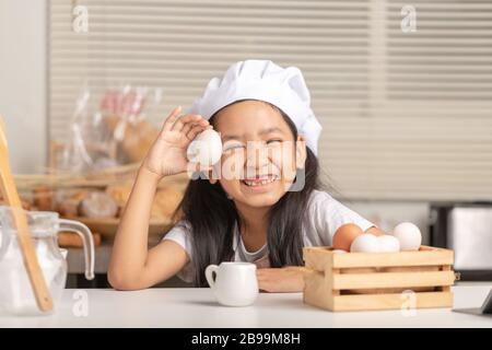 Asian little girl wearing a white chef hat is holding a duck egg and smiling. Eggs in a wooden basket in the kitchen. Stock Photo