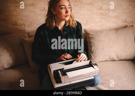 Young female writer using typewriter while sitting on sofa at home Stock Photo