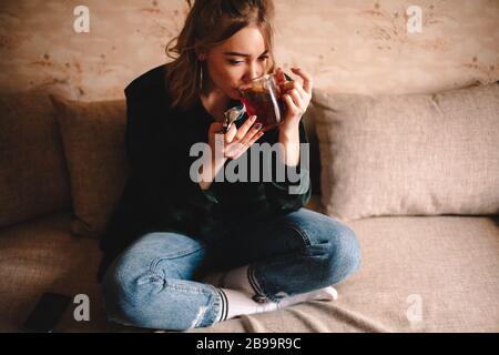 Young thoughtful woman drinking tea and eating chocolate while sitting on sofa at home Stock Photo