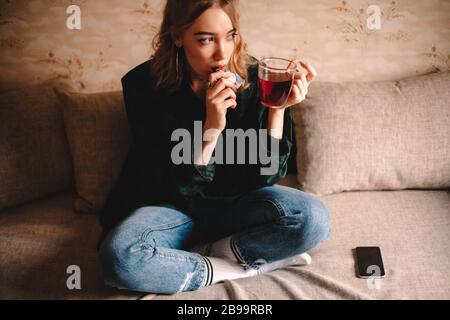 Young thoughtful woman drinking tea and eating chocolate while sitting on sofa at home Stock Photo