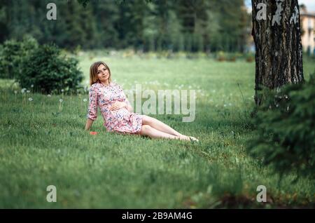 Pregnant woman inpink dress sitting on grass and touching bump whilst holding pink rose girl waiting Stock Photo