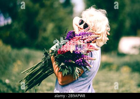 Blonde mature stout woman holding a lupine bouquet on nature background. The concept of beauty and Body Positive. Sunset light Stock Photo