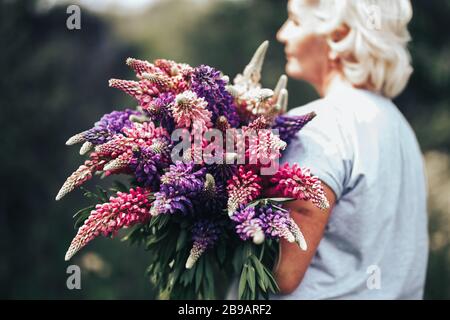 Blonde mature stout woman holding a lupine bouquet on nature background. The concept of beauty and Body Positive. Sunset light Stock Photo