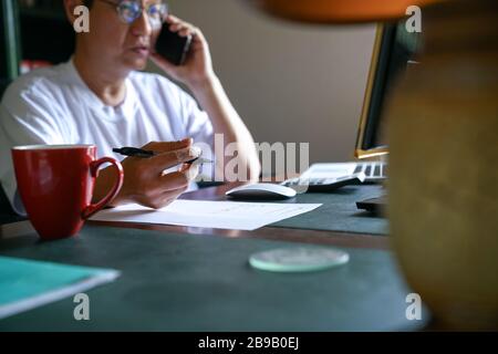 Man working from home, on his cell phone and facing computer screen.  Focus on his hand with pen. Home office concept. Stock Photo