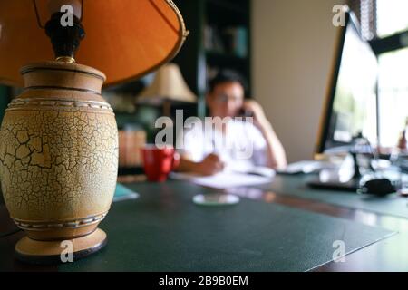 Work from home concept. Focus on table lamp with blurred home background and man on cell phone. Stock Photo