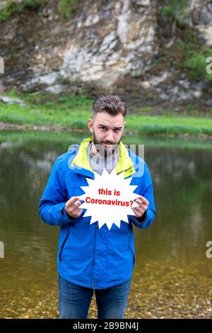Portrait of handsome young man in jeans and blue sport jacket on bank of clear mountain lake with cartoon star plate asking if this is coronavirus in Stock Photo