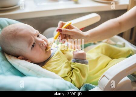 Mom feeds baby vegetable puree in a high chair. The mother puts a spoon of mashed in the child's mouth. The infant eats food. Stock Photo