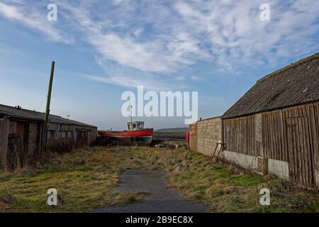 An old traditional wooden Fishing boat up in blocks and out of the water between old wooden and derelict sheds and garages. Stock Photo