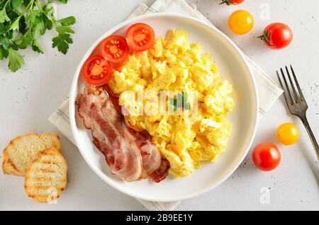 Scrambled eggs, fried bacon and tomatoes on plate over light stone background. Top view, flat lay Stock Photo