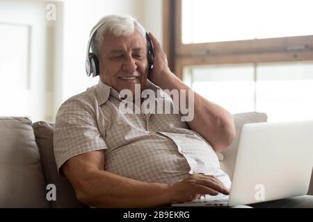 Smiling older man wearing wireless headphones enjoying music, using laptop Stock Photo