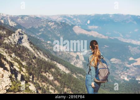 Young beautiful girl travels alone in the mountains in spring or autumn, looks into the distance and enjoys nature, rocks and green forests, view of the landscape. a backpack behind and sportswear, freedom and lightness Stock Photo