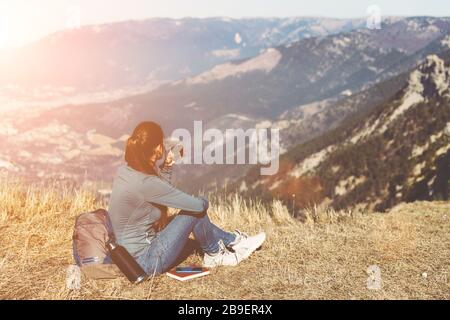 Young beautiful girl travels alone in the mountains in spring or autumn, sits on the edge of the mountain and looks into the distance and enjoys nature, rocks and green forests, view of the landscape. a backpack behind and sportswear, a thermos with a hot drink or tea, freedom and lightness. Back view of woman traveler in cap sitting on mountain top alone and looking at beautiful summer landscape and blue sea view. Drink tea or coffe Stock Photo