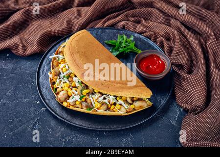 Malaysian street food apam balik - thick pancake stuffed with mushrooms, corn, shredded white cheese on a black plate with ketchup and parsley on a da Stock Photo