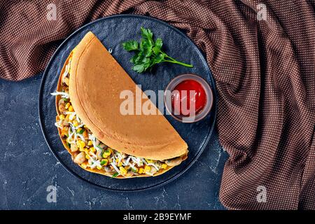 Malaysian apam balik - thick pancake stuffed with roasted mushrooms, corn, shredded white cheese on a black plate with ketchup and parsley on a dark c Stock Photo