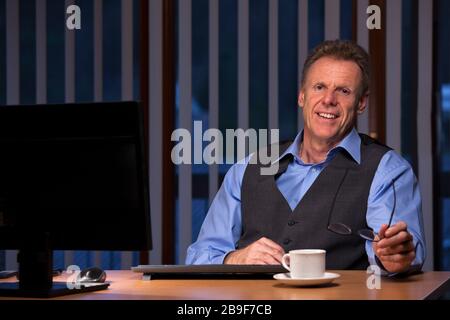 Senior businessman sitting at a desk with computer monitor in a dark blue office by night - smiling at the camera Stock Photo