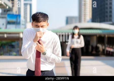 Asian ill businessman cough with mask with businesswoman in background to keep distance protect from COVID-19 viruses and people social distancing  fo Stock Photo