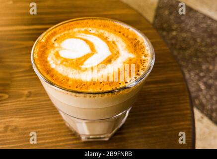A glass cup of milk coffee stands on the table, the view from above. A beautiful symmetrical pattern is made on coffee and a fragment of the mosaic floor is visible. Stock Photo