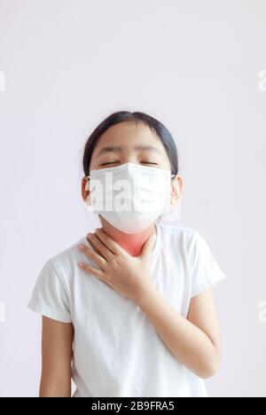 Little Asian girl touching her neck with a sore throat. The kid wearing a protective medical mask. The protection for the epidemic of the flu, Coronav Stock Photo