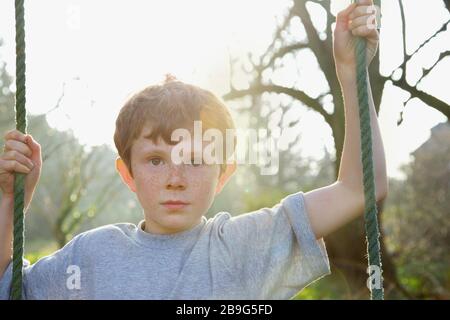 Portrait boy with freckles on rope swing in sunny garden Stock Photo