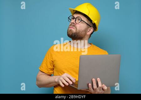 Male foreman or engineer in a construction helmet with a laptop, looking up seriously and displeasedly, on a blue background. Stock Photo