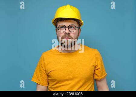 Puzzled bearded man in a construction helmet and round glasses looks up thoughtfully, on a blue background. Stock Photo