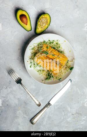 Cooked sweet corn with green alfalfa sprouts on plate and avocado and silver fork and knife on white table. Summer vegan dinner or snack. Stock Photo